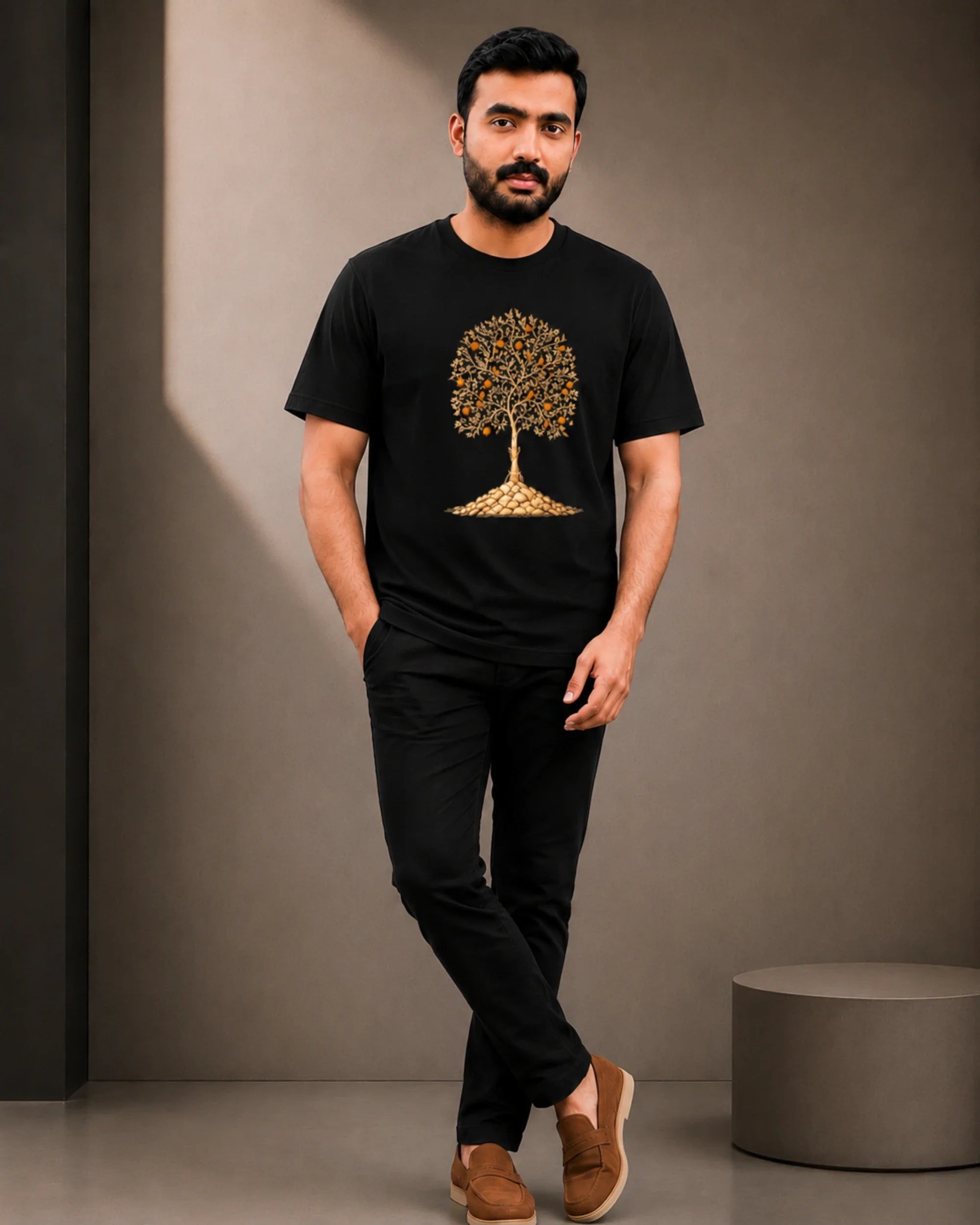 A man wearing a black short-sleeve t-shirt with a gold tree graphic on the front, paired with black pants and brown slip-on shoes, standing in a studio setting.