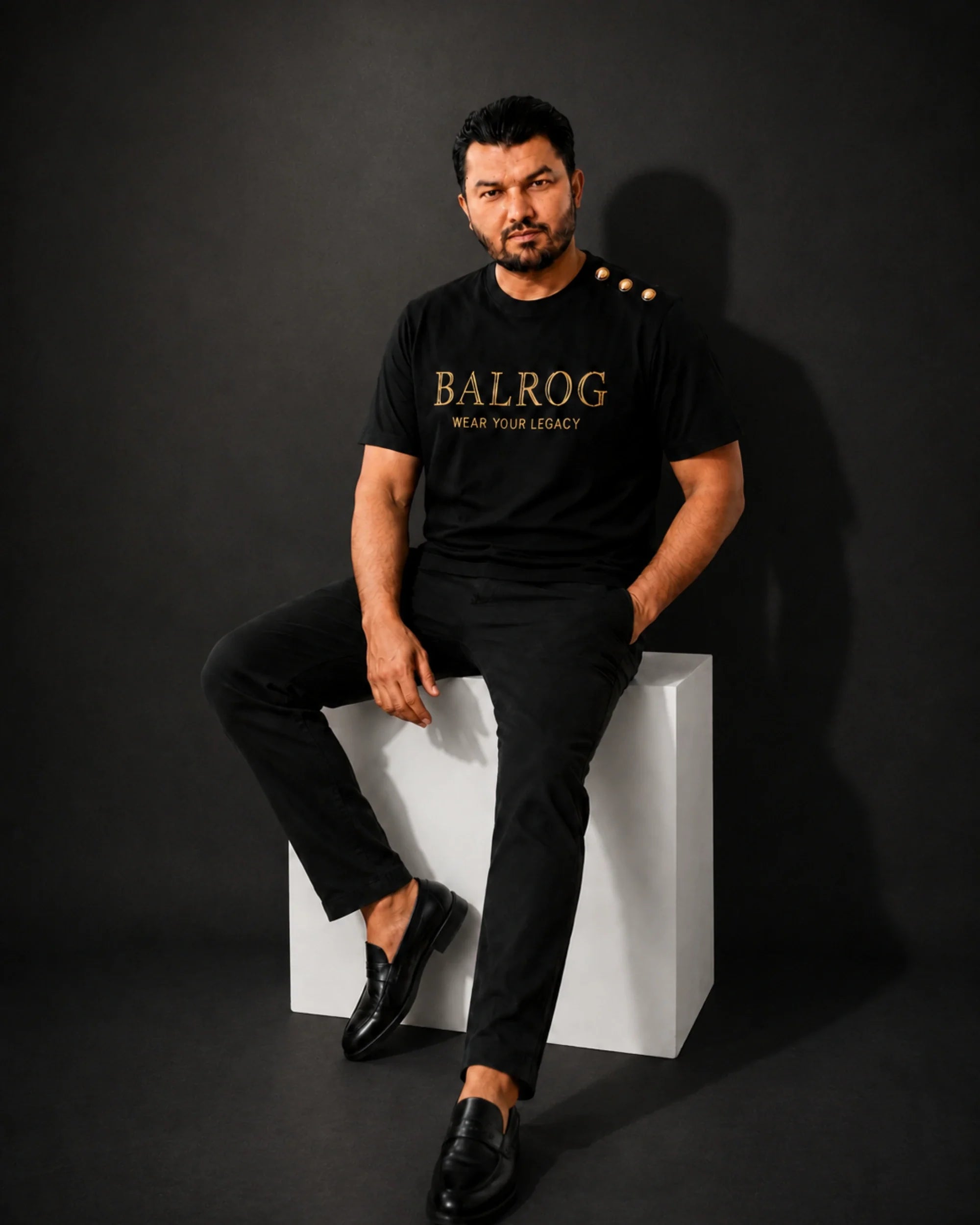 A man modeling a black Balrog t-shirt with gold lettering and three gold buttons on the shoulder, paired with black trousers, in a minimalist studio setting.