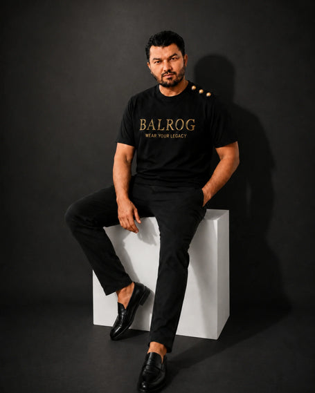 A man modeling a black Balrog t-shirt with gold lettering and three gold buttons on the shoulder, paired with black trousers, in a minimalist studio setting.