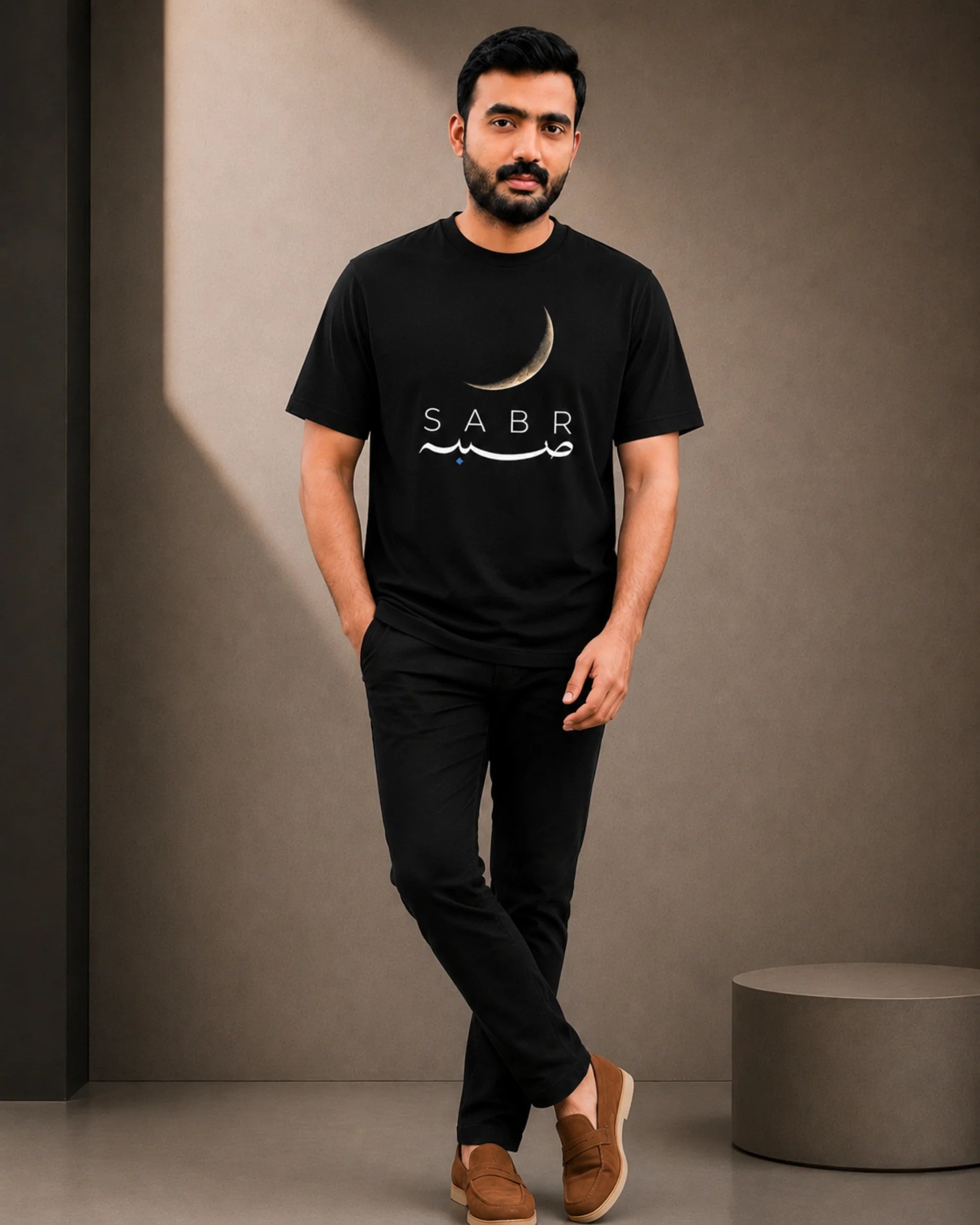 A man wearing a black crew-neck t-shirt with a white crescent moon and 'SABR' graphic, paired with black pants and brown slip-on shoes, standing against a textured gray wall.