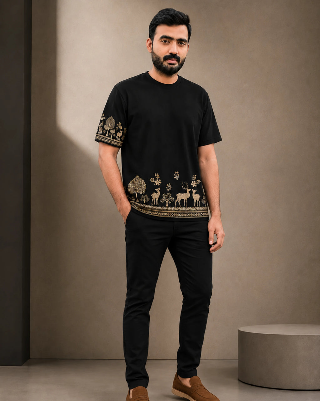 A man wearing a black short-sleeve t-shirt with beige and gold deer and tree patterns along the hem and sleeve cuffs, paired with black pants and brown shoes, standing against a textured beige wall.