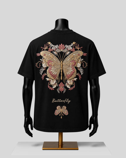 A black short-sleeve T-shirt displayed on a black mannequin torso with a gold stand, featuring a large ornate butterfly and floral graphic on the back in gold, pink, and beige tones, with the word 'Butterfly' printed below.