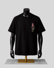 A black short-sleeve crewneck T-shirt displayed on a black mannequin torso with arms, featuring a small embroidered pink tulip on the left chest, set against a light gray background.