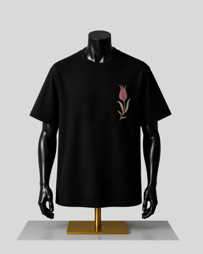 A black short-sleeve crewneck T-shirt displayed on a black mannequin torso with arms, featuring a small embroidered pink tulip on the left chest, set against a light gray background.