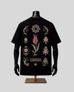 A black short-sleeve T-shirt displayed on a black mannequin torso with a gold stand, featuring ornate embroidered tulip and geometric motifs in beige and pink on the back, set against a solid light gray background.