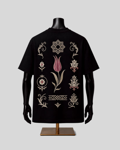 A black short-sleeve T-shirt displayed on a black mannequin torso with a gold stand, featuring ornate embroidered tulip and geometric motifs in beige and pink on the back, set against a solid light gray background.