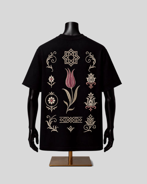 A black short-sleeve T-shirt displayed on a black mannequin torso with a gold stand, featuring ornate embroidered tulip and geometric motifs in beige and pink on the back, set against a solid light gray background.