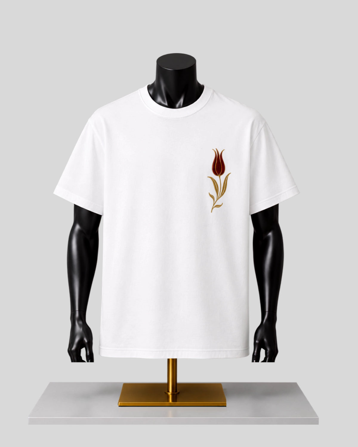 A white short-sleeve crewneck T-shirt displayed on a black mannequin torso with a gold stand, featuring a small embroidered tulip graphic in brown and gold on the left chest.