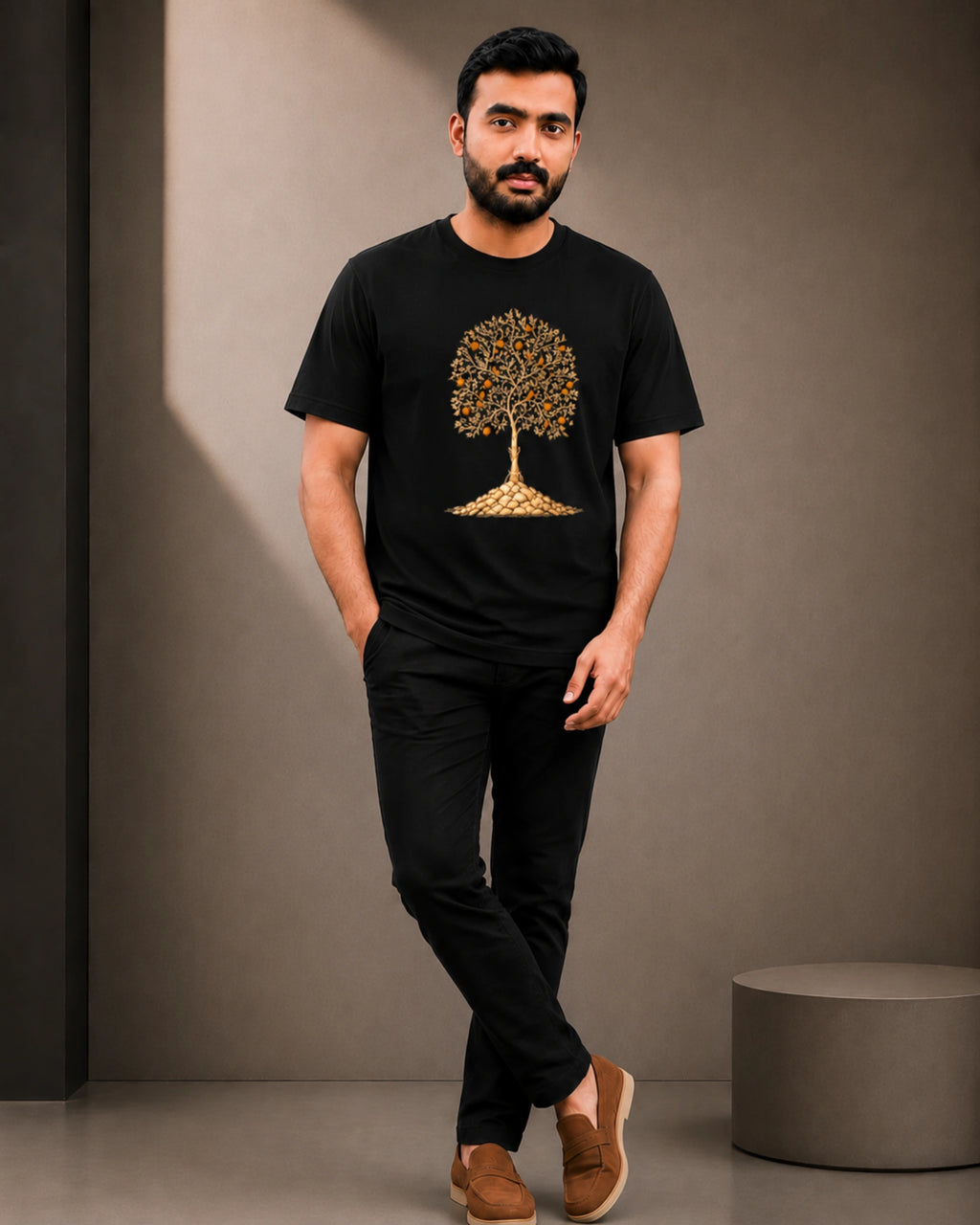 A man wearing a black short-sleeve t-shirt with a gold tree graphic on the front, paired with black pants and brown slip-on shoes, standing in a studio setting.