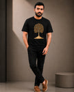 A man wearing a black short-sleeve t-shirt with a gold tree graphic on the front, paired with black pants and brown slip-on shoes, standing in a studio setting.