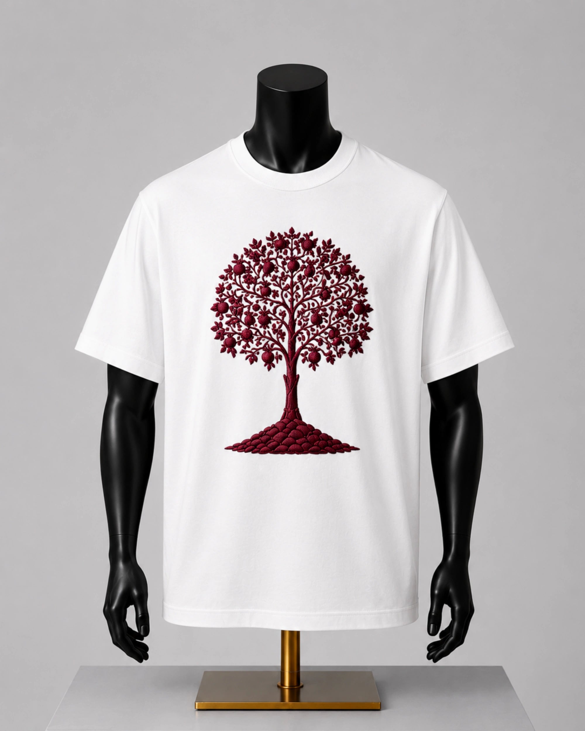 A white short-sleeve crewneck t-shirt displayed on a black mannequin torso with a gold stand, featuring a graphic of a tree made of red maple leaves and trunk.