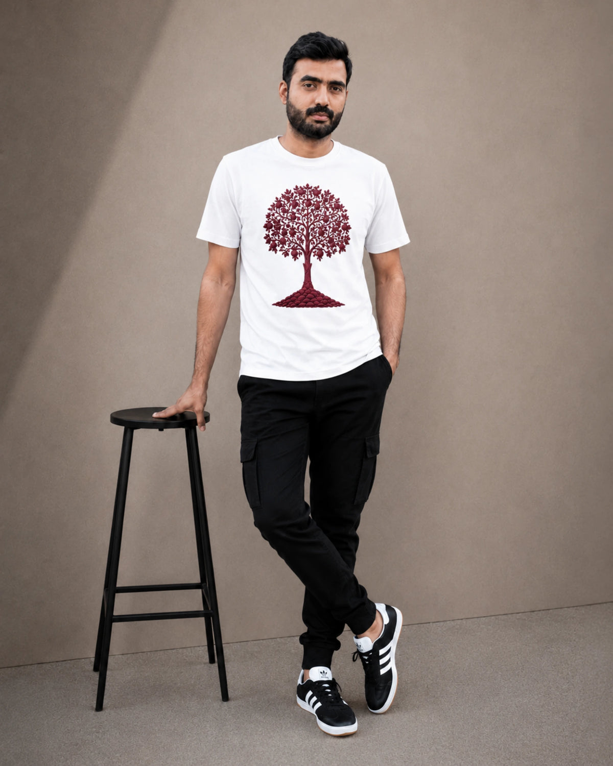 A man wearing a white crew-neck t-shirt with a red tree graphic, black cargo pants, and black-and-white sneakers, standing next to a black stool against a neutral beige studio background.