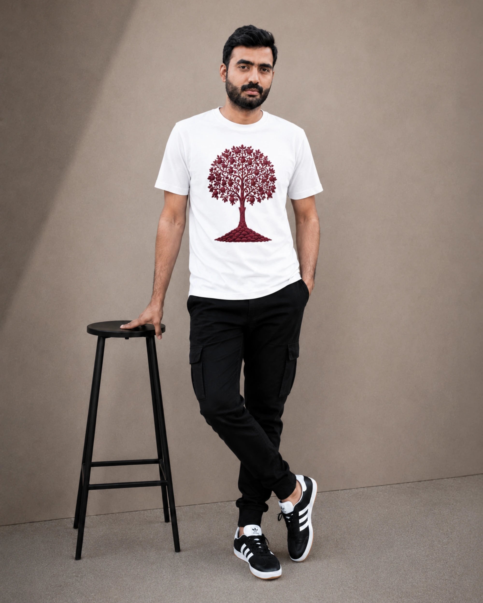 A man wearing a white crew-neck t-shirt with a red tree graphic, black cargo pants, and black-and-white sneakers, standing next to a black stool against a neutral beige studio background.