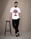 A man wearing a white crew-neck t-shirt with a red tree graphic, black cargo pants, and black-and-white sneakers, standing next to a black stool against a neutral beige studio background.