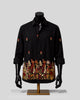 A black long-sleeve button-up shirt displayed on a black mannequin torso with arms. The shirt features small embroidered floral motifs on the chest and sleeves, and a large, dense floral and botanical border embroidery across the lower hem in red, gold, and cream tones.