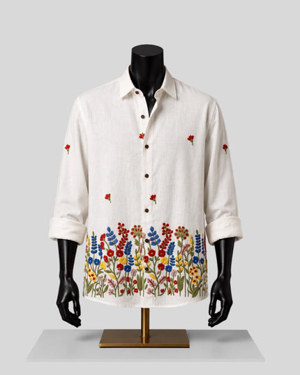 An off-white linen button-up shirt with colorful floral embroidery along the hem and scattered floral motifs on the chest and sleeves, displayed on a black mannequin torso with a gold stand against a light gray background.