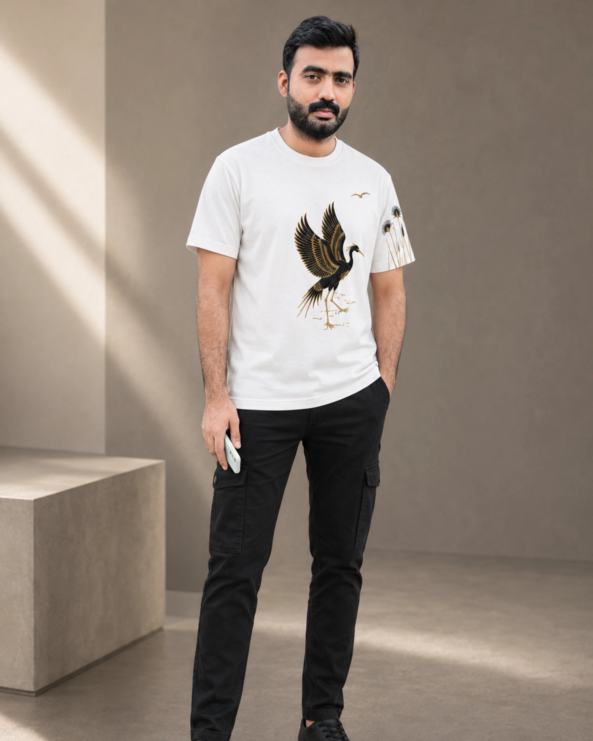 A man wearing a white crewneck t-shirt with a detailed graphic of a heron and a bird on the sleeve, paired with black cargo pants, standing in a minimalist studio setting.