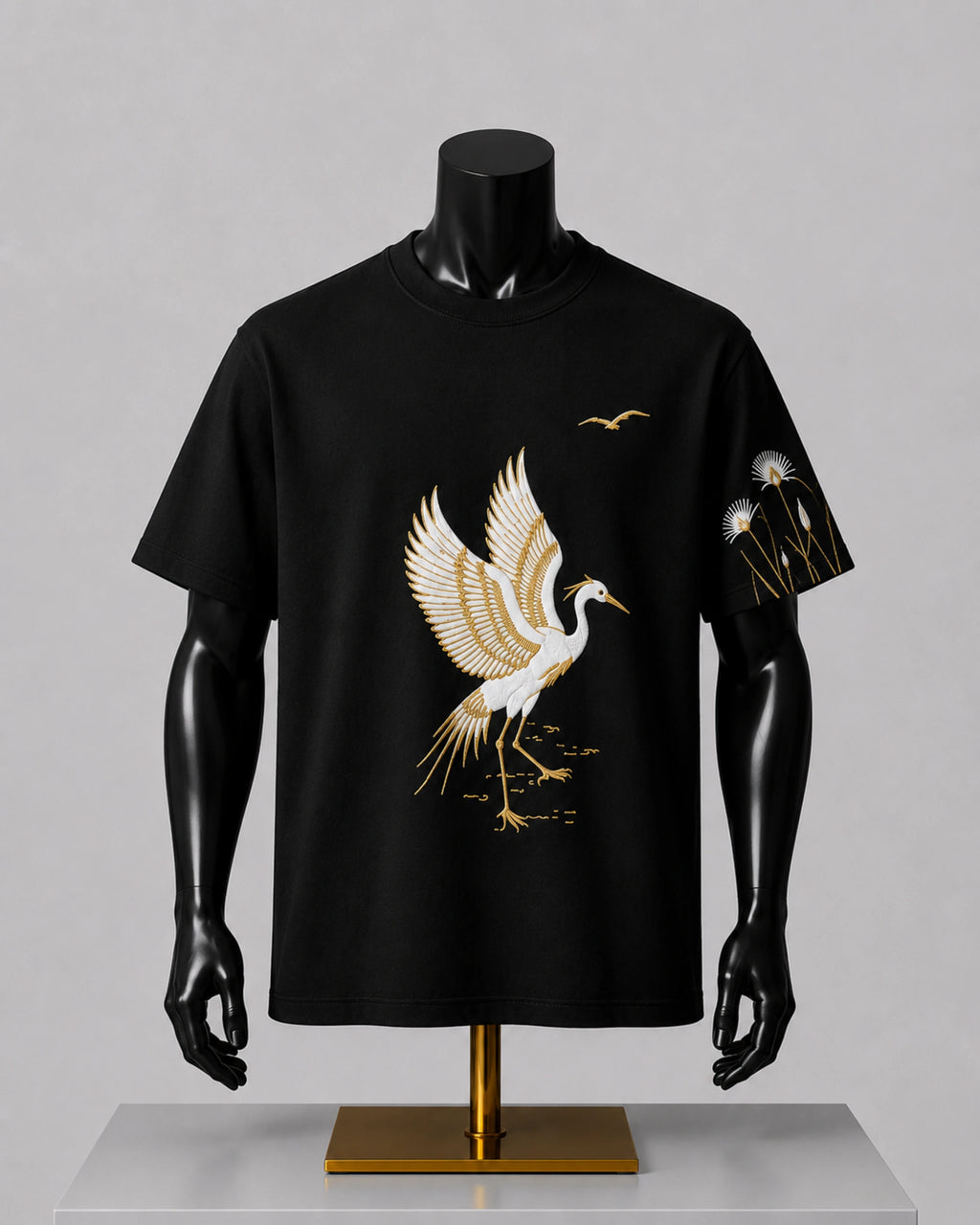 A black short-sleeve t-shirt displayed on a black mannequin torso with a gold stand, featuring a large white and gold crane graphic on the front and a smaller matching graphic on the sleeve.