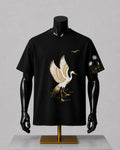 A black short-sleeve t-shirt displayed on a black mannequin torso with a gold stand, featuring a large white and gold crane graphic on the front and a smaller matching graphic on the sleeve.