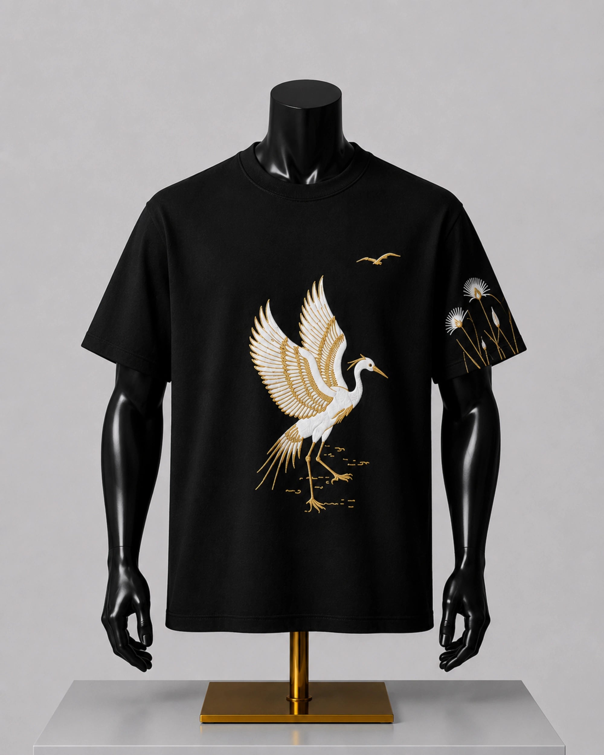 A black short-sleeve t-shirt displayed on a black mannequin torso with a gold stand, featuring a large white and gold crane graphic on the front and a smaller matching graphic on the sleeve.