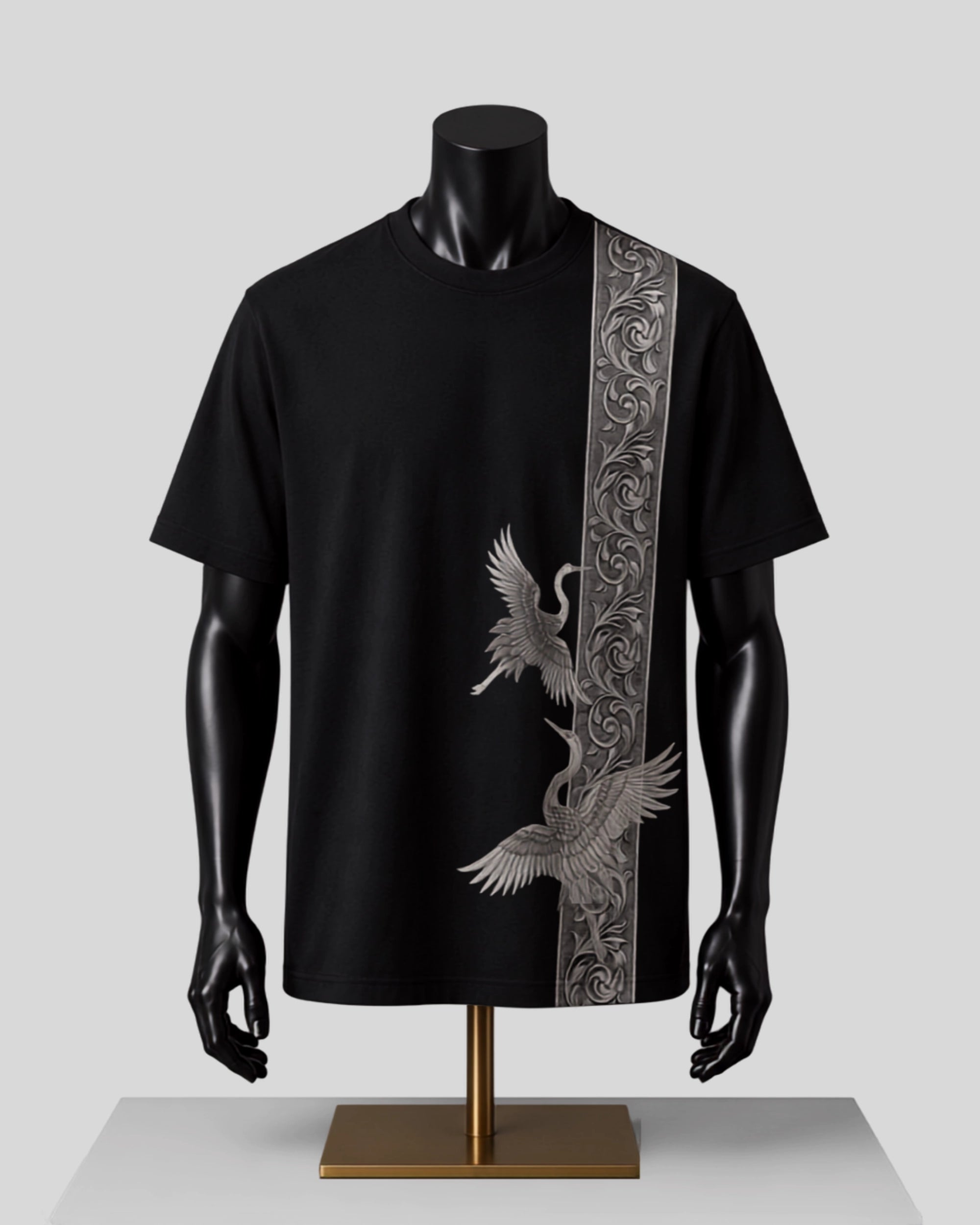 A black short-sleeve t-shirt displayed on a black mannequin torso with a gold stand, featuring a vertical silver-gray ornate pattern on the right side and two matching cranes on the front.
