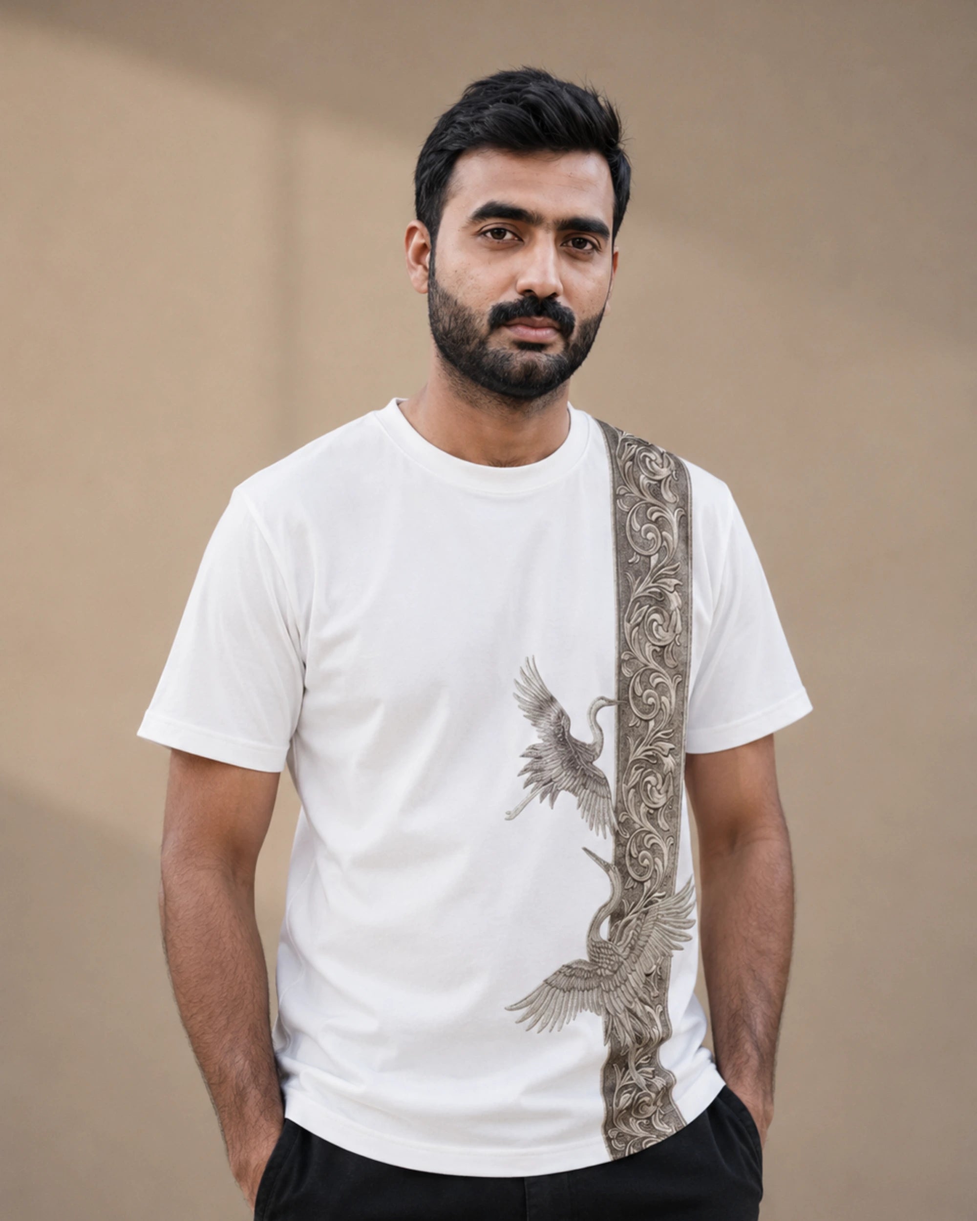 A man wearing a white crew-neck t-shirt with a vertical brown and beige ornate pattern on the right side and a crane graphic, paired with black pants, against a plain beige background.
