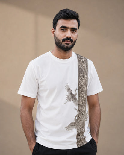 A man wearing a white crew-neck t-shirt with a vertical brown and beige ornate pattern on the right side and a crane graphic, paired with black pants, against a plain beige background.
