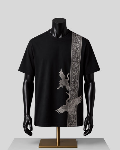 A black short-sleeve t-shirt displayed on a black mannequin torso with a gold stand, featuring a vertical silver-gray ornate patterned stripe down the right side and a large silver crane graphic on the front.