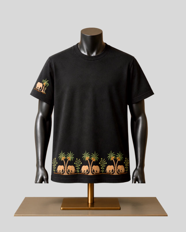 A black short-sleeve t-shirt displayed on a black mannequin torso, featuring a graphic print of brown elephants and green palm trees along the bottom hem and left sleeve, set against a light gray background.