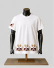 A white short-sleeve crewneck t-shirt displayed on a black mannequin torso over a gold stand. The shirt features a repeating border print of brown elephants and gold palm trees along the hem, and a matching motif on the left sleeve.