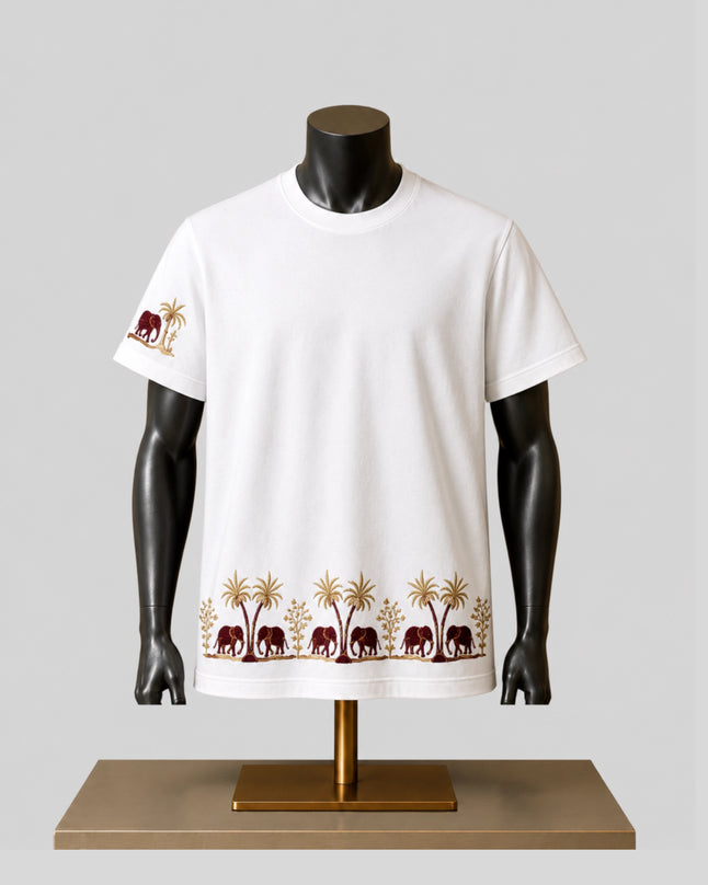 A white short-sleeve crewneck t-shirt displayed on a black mannequin torso over a gold stand. The shirt features a repeating border print of brown elephants and gold palm trees along the hem, and a matching motif on the left sleeve.