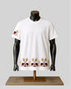 A white short-sleeve crewneck t-shirt displayed on a black mannequin torso over a gold stand. The shirt features a repeating border print of brown elephants and gold palm trees along the hem, and a matching motif on the left sleeve.