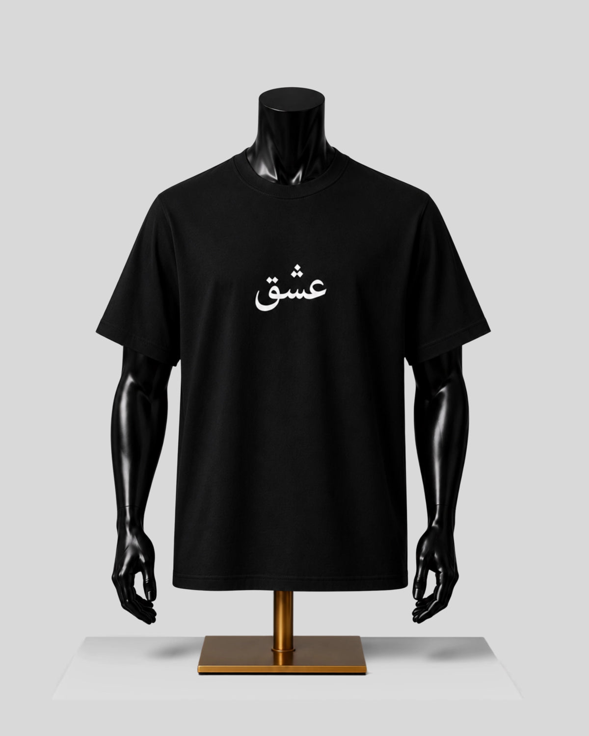 A black short-sleeve crewneck t-shirt displayed on a black mannequin torso with a gold stand, featuring white Arabic calligraphy printed on the chest, set against a light gray background.