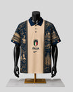 A short-sleeve polo shirt displayed on a black mannequin torso with a gold stand against a light gray background. The shirt has a beige center panel with the Italian crest and the word 'ITALIA' above a Nike swoosh, flanked by dark navy blue panels featuring architectural and historical illustrations.