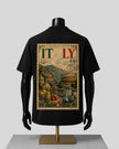 A black short-sleeve T-shirt displayed on a black mannequin torso with a gold stand, shown from the back. The shirt features a large vintage-style Italy travel stamp graphic with illustrations of the Colosseum, mountains, and Italian landscapes, along with the word 'ITALY' and a postmark.