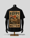 A black short-sleeve T-shirt displayed on a black mannequin torso, showing the back side with a large vintage-style Egyptian travel poster graphic. The graphic includes the text 'PROUD TO BE EGYPT' and an illustrated scene with the Egyptian flag, pyramids, and desert landscape in beige, brown, red, and green tones.