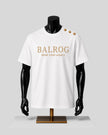 White short-sleeve T-shirt displayed on a black mannequin torso with gold text reading 'BALROG WEAR YOUR LEGACY' and three gold buttons near the right shoulder, set against a light gray background.
