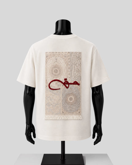 An off-white short-sleeve T-shirt displayed on a black mannequin torso, showing the back side. The shirt features a large rectangular graphic print of a beige and gray patterned rug with a bold red Arabic calligraphy design across the center.