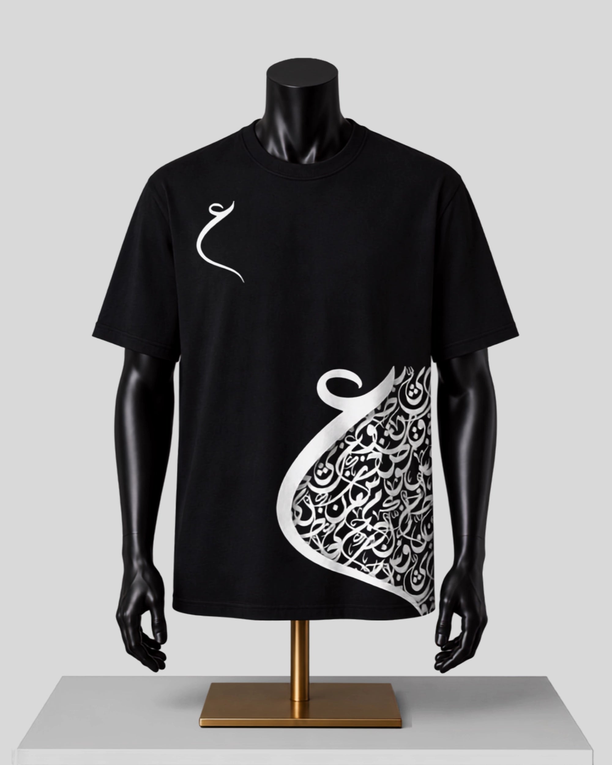 A black short-sleeve t-shirt displayed on a black mannequin torso with a gold stand against a light gray background. The shirt features a white Arabic calligraphy graphic on the right side and a small white calligraphy motif on the left chest.