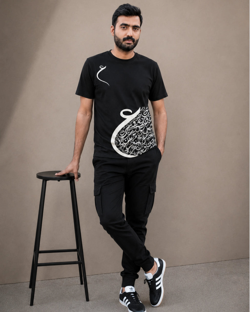A man wearing a black short-sleeve t-shirt with a white Arabic calligraphy graphic, black cargo pants, and black sneakers, standing next to a black stool against a plain beige background.