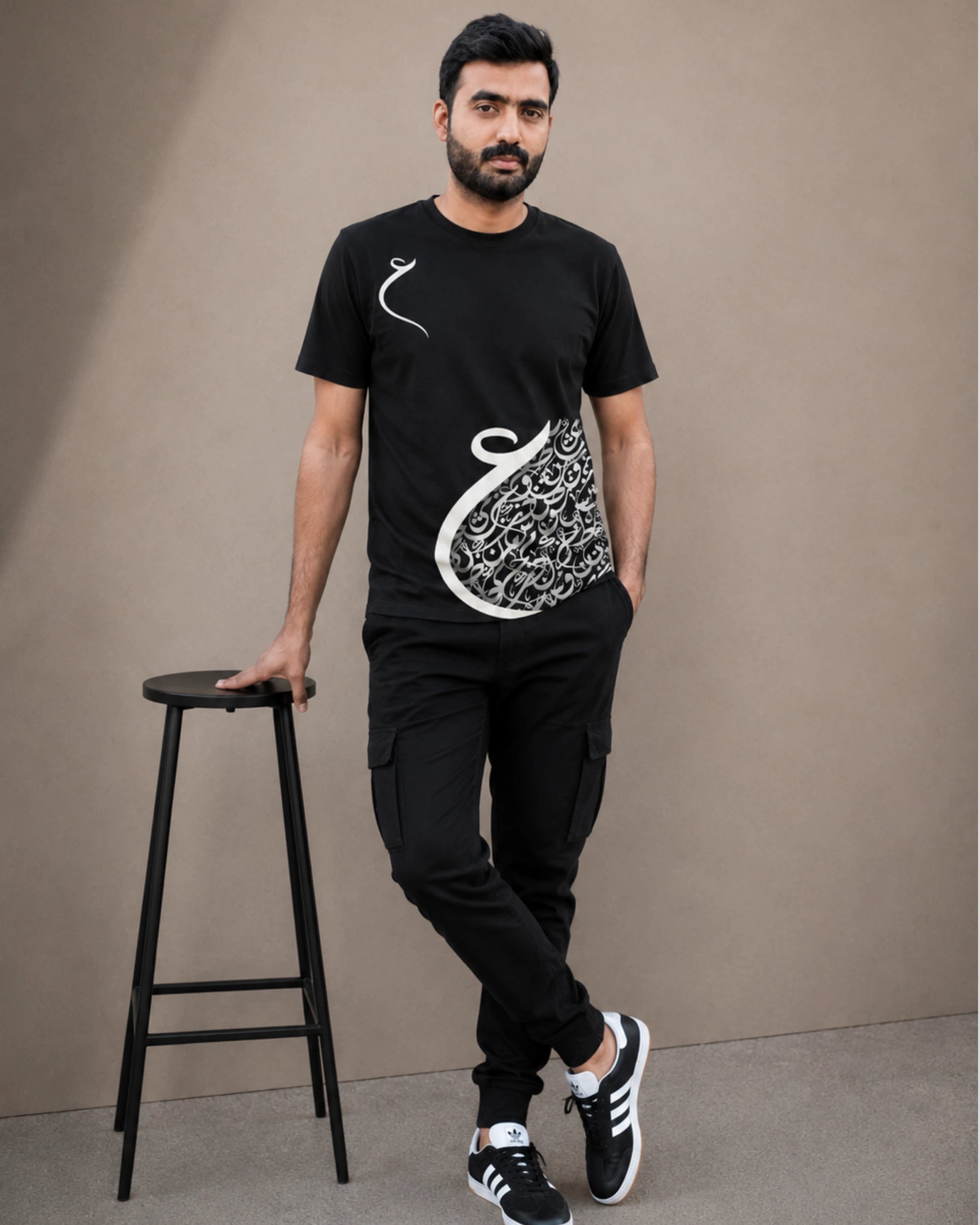 A man wearing a black short-sleeve t-shirt with a white Arabic calligraphy graphic, black cargo pants, and black sneakers, standing next to a black stool against a plain beige background.