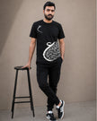 A man wearing a black short-sleeve t-shirt with a white Arabic calligraphy graphic, black cargo pants, and black sneakers, standing next to a black stool against a plain beige background.