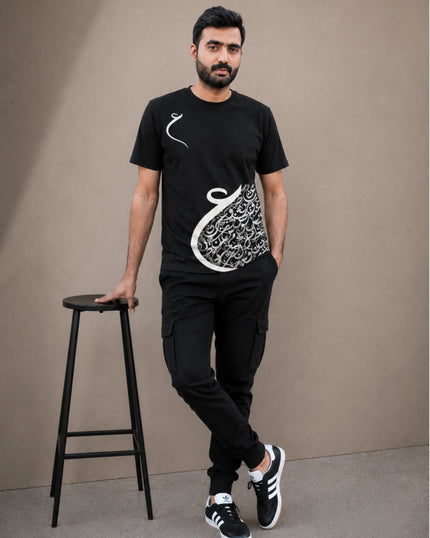 A man wearing a black short-sleeve t-shirt with a white Arabic calligraphy graphic, black cargo pants, and black sneakers, standing next to a black stool against a plain beige background.