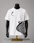 A white short-sleeve t-shirt displayed on a black mannequin torso with a gold stand against a neutral gray background. The shirt features a black Arabic calligraphy graphic, resembling a stylized crescent or abstract scroll, printed on the right side and a small matching calligraphy motif on the left chest.