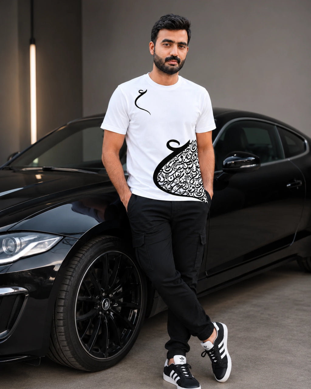 A man wearing a white graphic t-shirt with a black abstract design, black pants, and black sneakers, leaning against a black sports car in a modern indoor setting.