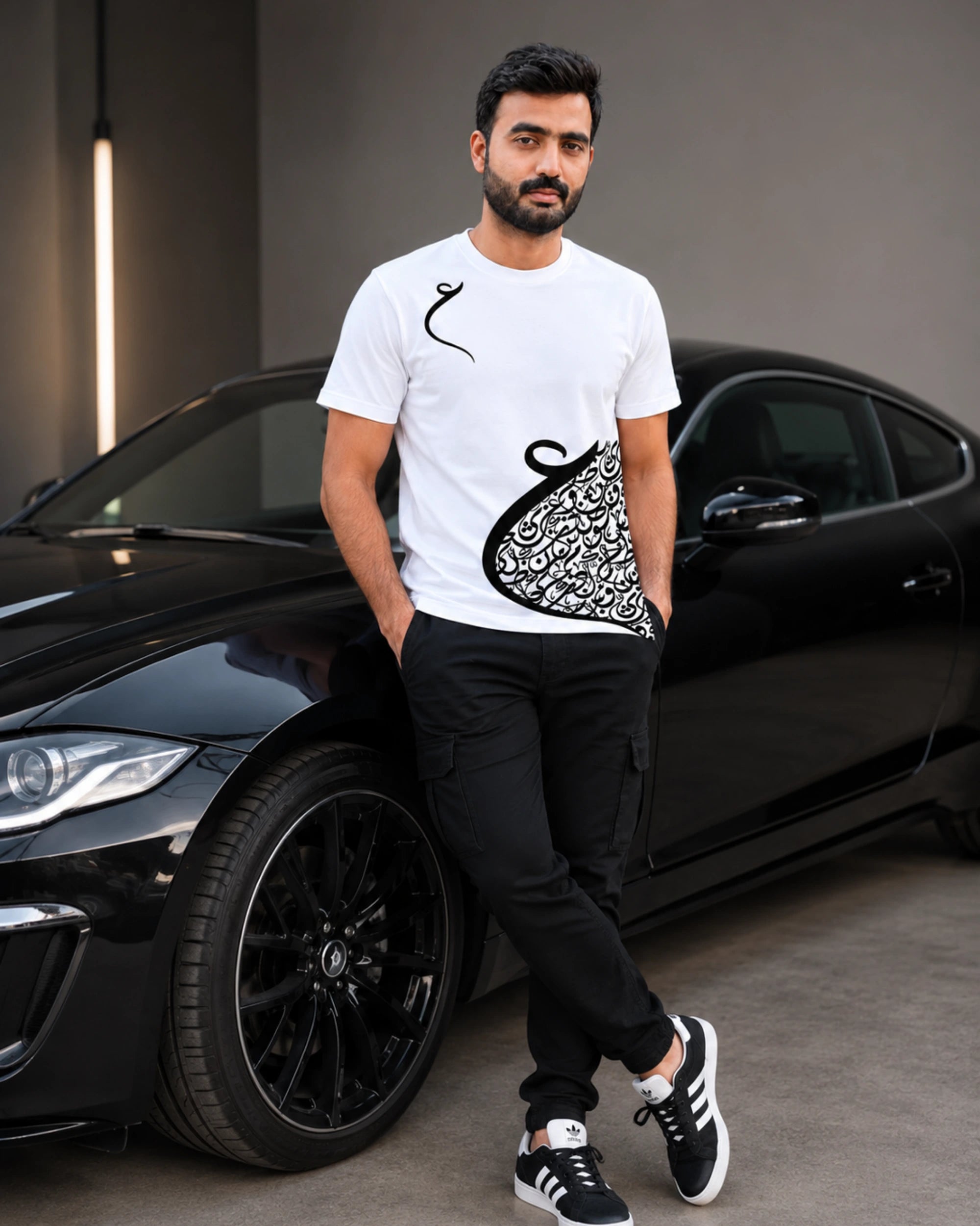 A man wearing a white graphic t-shirt with a black abstract design, black pants, and black sneakers, leaning against a black sports car in a modern indoor setting.