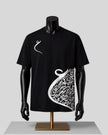 A black short-sleeve t-shirt displayed on a black mannequin torso with a gold stand against a light gray background. The shirt features a white Arabic calligraphy graphic on the right side and a small white calligraphy symbol on the left chest.