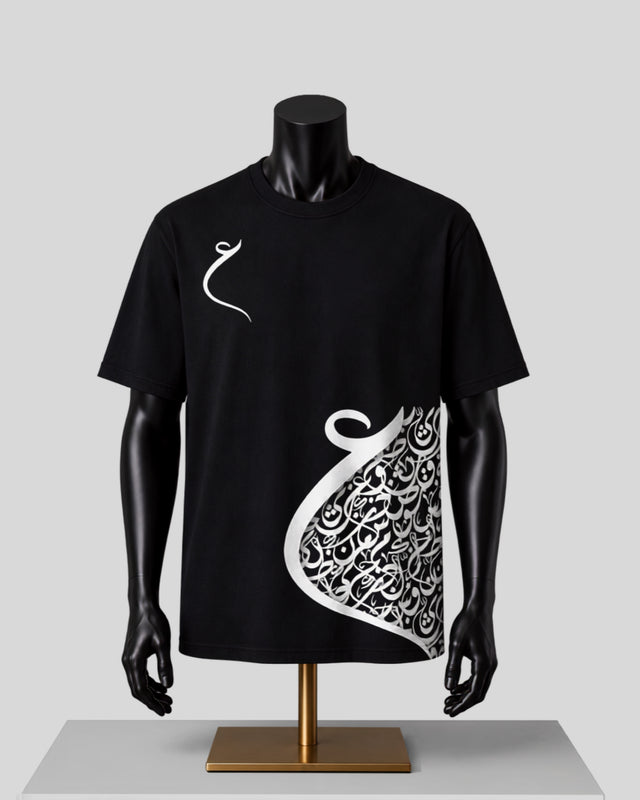 A black short-sleeve t-shirt displayed on a black mannequin torso with a gold stand against a light gray background. The shirt features a white Arabic calligraphy graphic on the right side and a small white calligraphy symbol on the left chest.