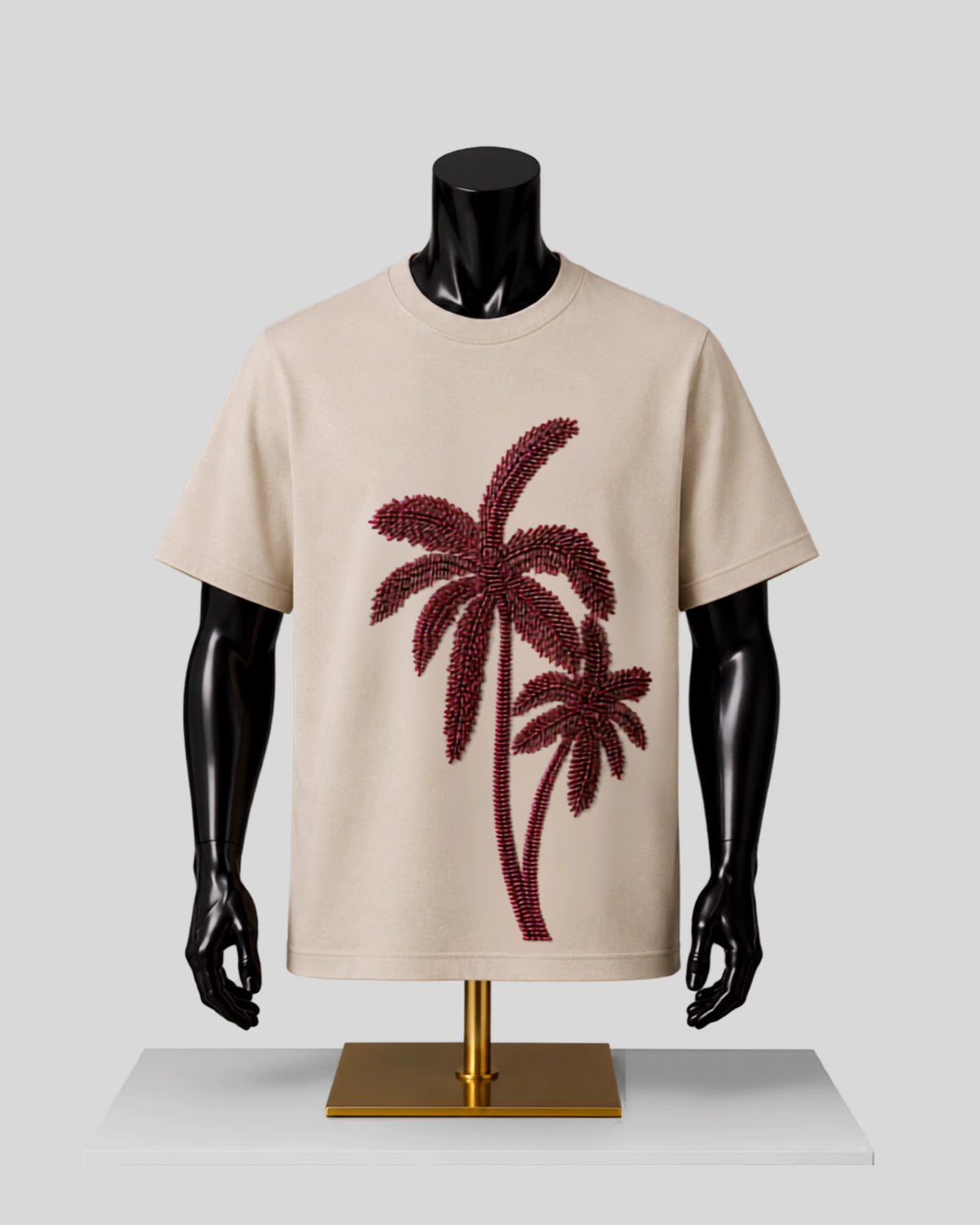 A beige short-sleeve crewneck T-shirt displayed on a black mannequin torso with a gold stand, featuring a large dark red palm tree graphic on the front.