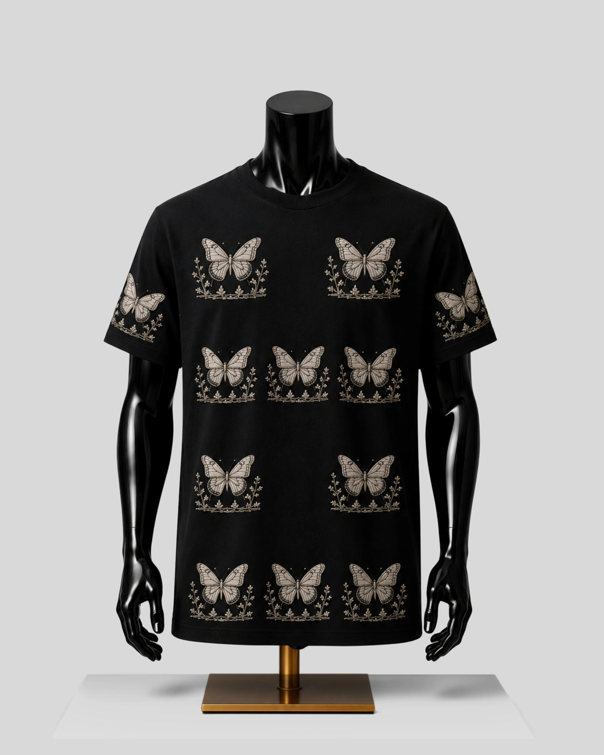 A black short-sleeve t-shirt with a repeating beige butterfly and floral pattern, displayed on a black mannequin torso with a gold stand against a light gray background.